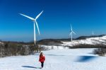 Beautiful Girl Walking In Winter Landscape Of Sky And Winter Road With Snow And Red Dress And Wind Turbine Stock Photo