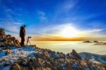 Professional Photographer Takes Photos With Camera On Tripod On Rocky Peak At Sunset Stock Photo