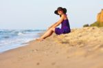 Cute White Girl In Hat And Purple Dress Sitting On Sand Stock Photo