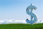 Damaged Concrete Dollar Sign Statue Isolated On Grass Meadow With Snowy Mountain And Blue Sky As Background Stock Photo