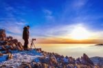 Professional Photographer Takes Photos With Camera On Tripod On Rocky Peak At Sunset Stock Photo