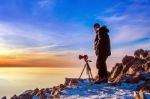 Professional Photographer Takes Photos With Camera On Tripod On Rocky Peak At Sunset Stock Photo