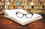 Open Book With Glasses On The Desk Against Library Stock Photo