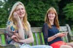 Two Girls Sitting On Bench In Park With Mobile Stock Photo