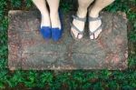 Top View Feet Of Male And Female Couple Lover Standing On Stone Floor Among Little Green Grass Garden. Hipster Style Stock Photo