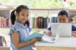 Group Of Children Reading, Discussing Interesting Book With Laptop Computer In Library Elementary School Stock Photo