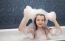 Little Girl Sitting In A Bath And Imposes Soap Suds On Her Head