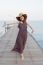 Portrait Of Beautiful Young Woman Wearing Wide Straw Hat And Long Dress Standing With Happiness Emotion On Piers At Sea Beach