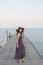 Portrait Of Beautiful Young Woman Wearing Wide Straw Hat And Long Dress Standing With Happiness Emotion On Piers At Sea Beach