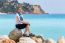 European Woman As Tourist Sitting On Rocks Near Blue Sea