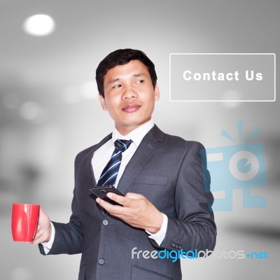 Asian Businessman With Cup Of Coffee And Using Smartphone Looking Away On Contact Us Sign Stock Photo