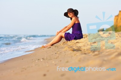 Cute White Girl In Hat And Purple Dress Sitting On Sand Stock Photo