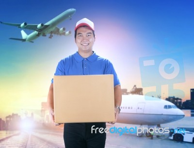 Delivery Man Holding Card Box Toothy Smiling Face Against Shipping Port And Cargo Plane Flying Background Stock Photo