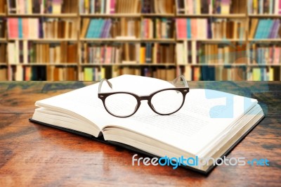 Open Book With Glasses On The Desk Against Library Stock Photo