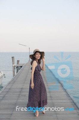 Portrait Of Beautiful Young Woman Wearing Wide Straw Hat And Long Dress Standing With Happiness Emotion On Piers At Sea Beach Stock Photo