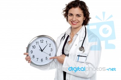 Pretty Doctor Pointing Out Time On Wall Clock Stock Photo