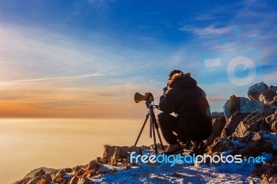 Professional Photographer Takes Photos With Camera On Tripod On Rocky Peak At Sunset Stock Photo