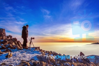 Professional Photographer Takes Photos With Camera On Tripod On Rocky Peak At Sunset Stock Photo