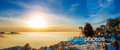 Professional Photographer Takes Photos With Camera On Tripod On Rocky Peak At Sunset Stock Photo