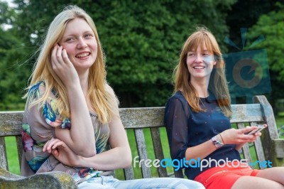 Two Girls Sitting On Bench In Park With Mobile Stock Photo