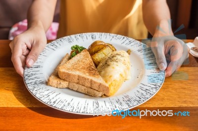 Woman Hand Holding White Plate With Omelet Potato, Tomatoes Parsley And Feta Cheese And Bread On Wooden Table. Eating Breakfast Stock Photo