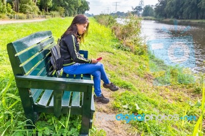 Woman Operating Mobile Phone On Bench At River Stock Photo