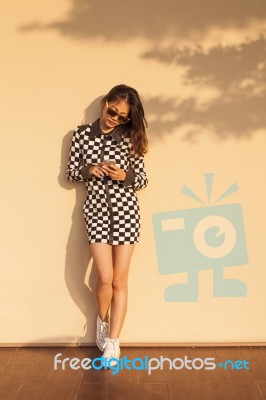 Young And Beautiful Hipster Woman Wearing Sun Glasses And Reading Social Media In Smart Phone Against Warm Yellow Wall At Home Terrace Stock Photo