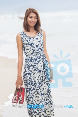 Young Asian Woman With Smiling Face Wearing Long Dress Standing With Sneaker In Hand On Sea Beach Stock Photo