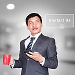 Asian Businessman With Cup Of Coffee And Using Smartphone Looking Away On Contact Us Sign Stock Photo