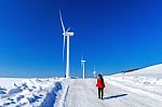Beautiful Girl Walking In Winter Landscape Of Sky And Winter Road With Snow And Red Dress And Wind Turbine Stock Photo